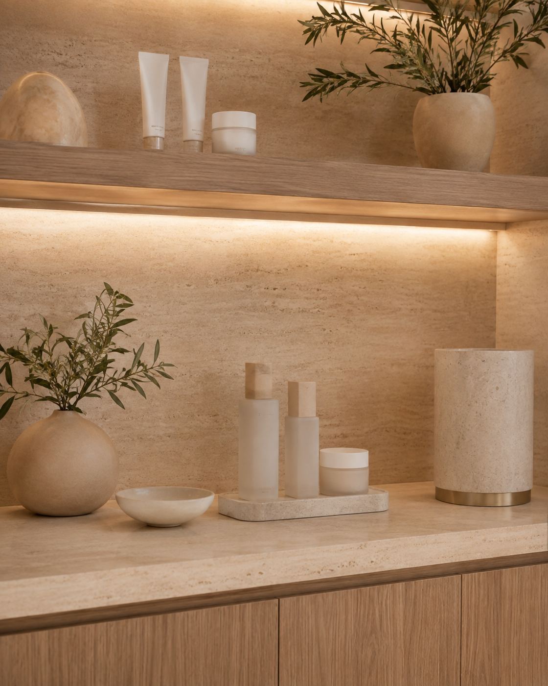 Bancada de travertino com produtos de skincare, vaso de cerâmica com ramos e nicho iluminado ao fundo.
